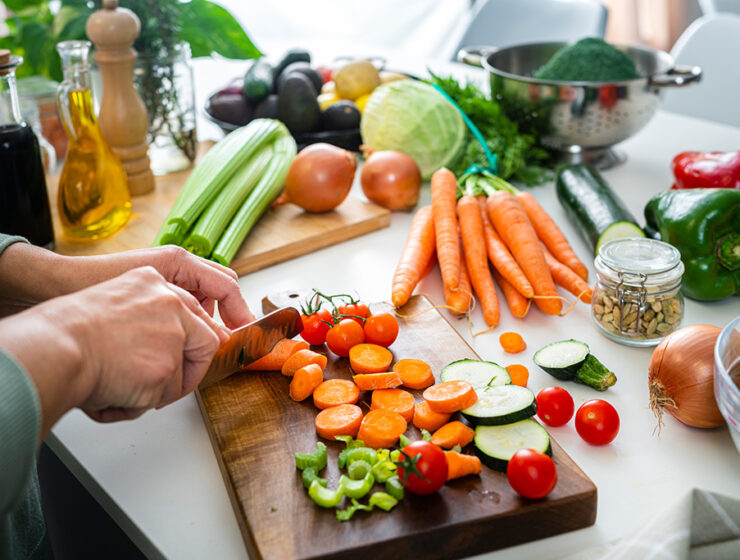 A person chops fresh vegetables on a wooden cutting board, surrounded by various colorful veggies, oils, and seasonings in a bright kitchen.