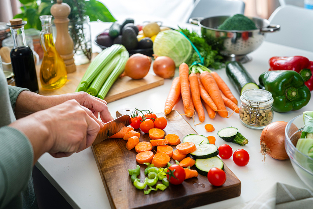 A person chops fresh vegetables on a wooden cutting board, surrounded by various colorful veggies, oils, and seasonings in a bright kitchen.