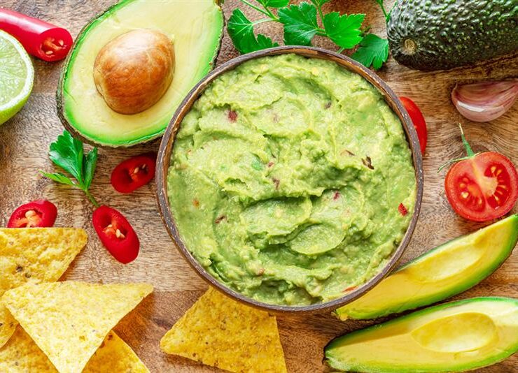 Bowl of fresh guacamole surrounded by ingredients like avocados, lime, cherry tomatoes, red chili peppers, and tortilla chips on a wooden surface.