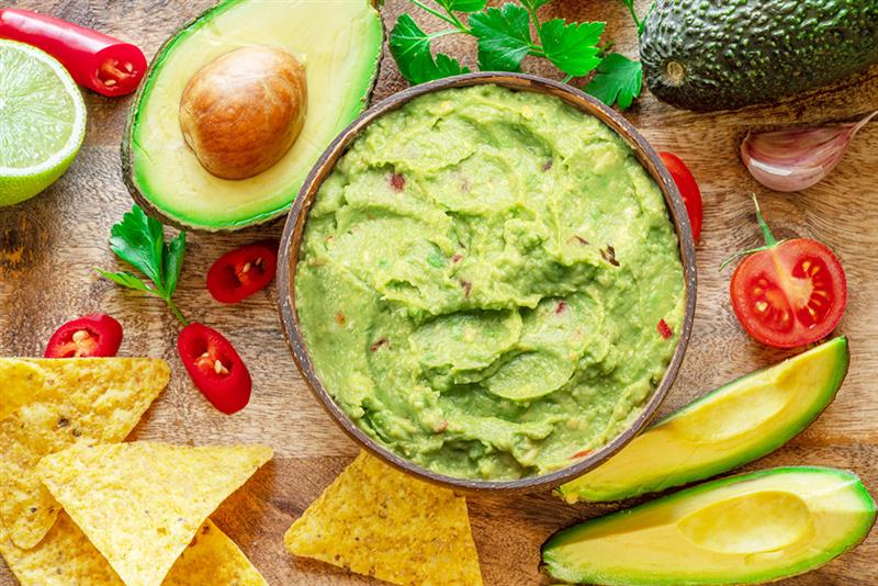 Bowl of fresh guacamole surrounded by ingredients like avocados, lime, cherry tomatoes, red chili peppers, and tortilla chips on a wooden surface.