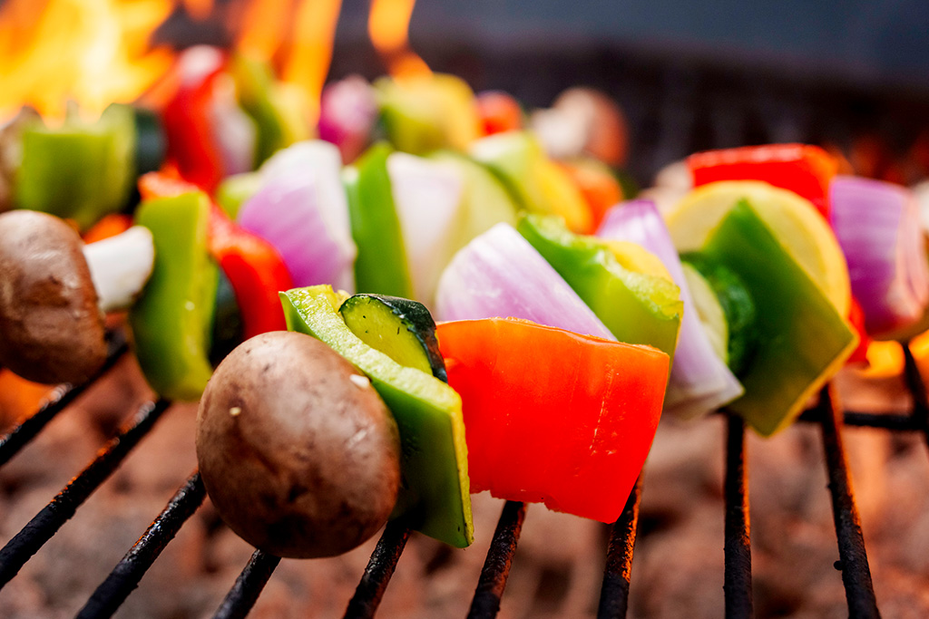 Close-up of colorful vegetable skewers with bell peppers, onions, zucchini, and mushrooms grilling over an open flame.