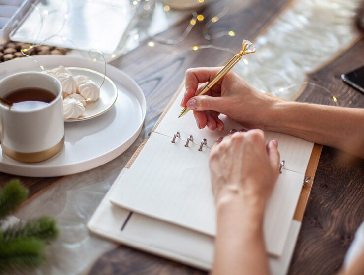 Person writing in a journal with a decorative pen, accompanied by a cup of tea and meringues on a table for a cozy setting.