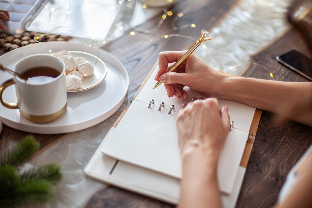 Person writing in a journal with a decorative pen, accompanied by a cup of tea and meringues on a table for a cozy setting.