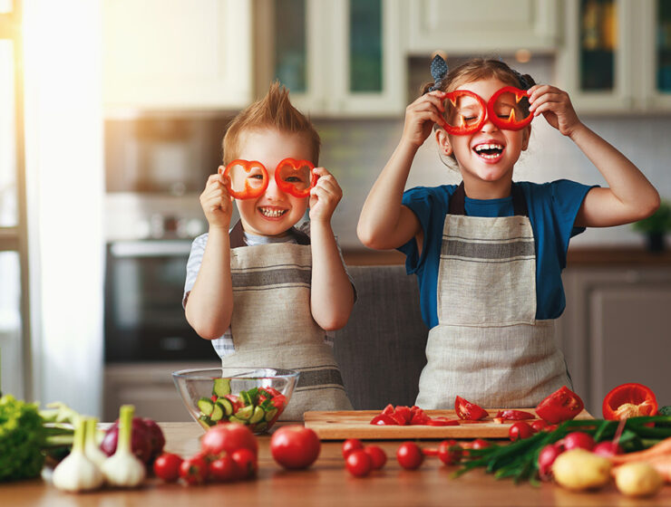 Two children in aprons having fun in the kitchen, holding bell pepper slices in front of their eyes, surrounded by fresh vegetables.