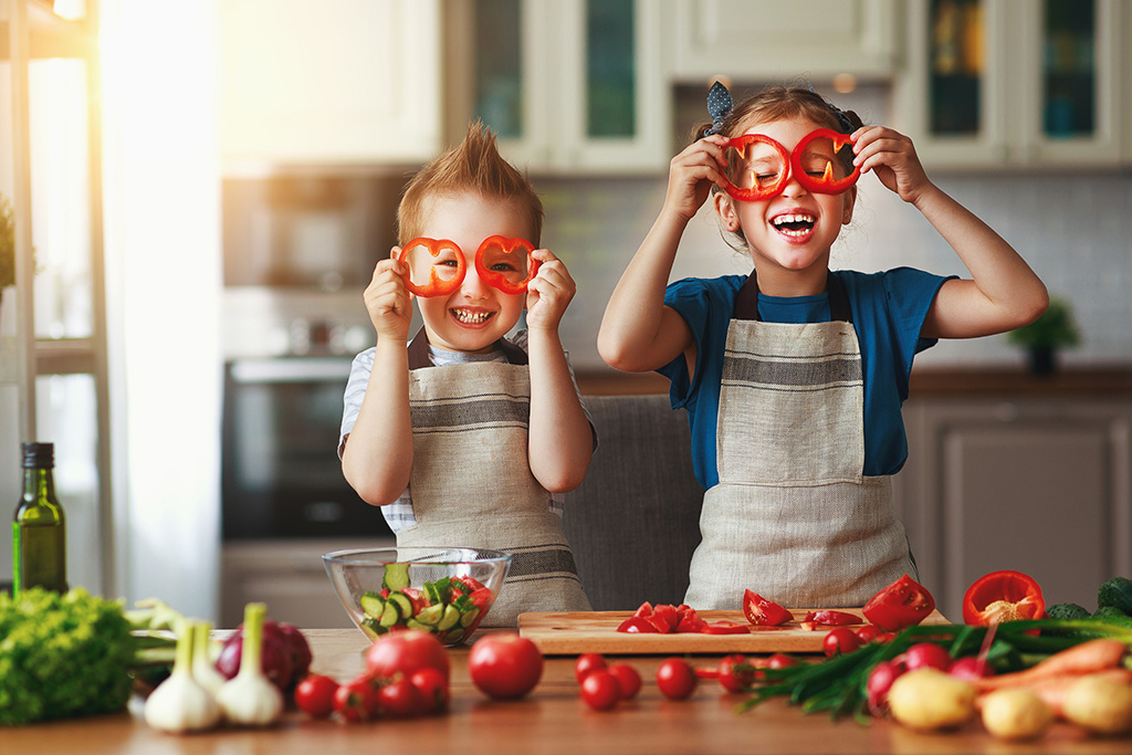 Two children in aprons having fun in the kitchen, holding bell pepper slices in front of their eyes, surrounded by fresh vegetables.