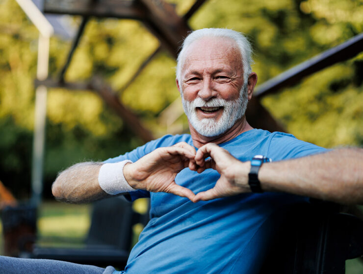 Happy elderly man in a park making a heart shape with his hands, wearing a blue shirt and wristband, enjoying an active lifestyle.