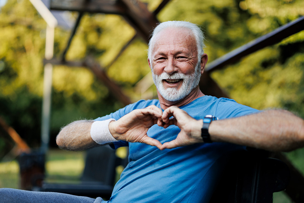 Happy elderly man in a park making a heart shape with his hands, wearing a blue shirt and wristband, enjoying an active lifestyle.