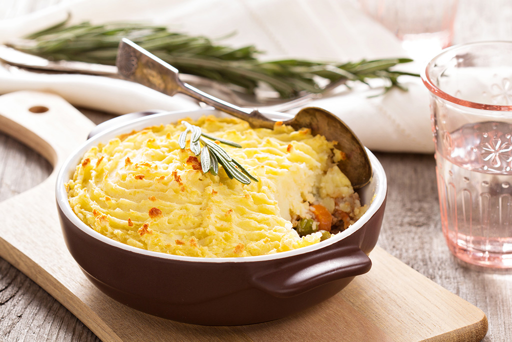 Shepherd's pie with golden mashed potato topping in a rustic dish, garnished with rosemary on a wooden board.
