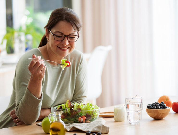 Smiling woman enjoying a fresh salad at a dining table, surrounded by healthy ingredients like fruits, vegetables, and water.