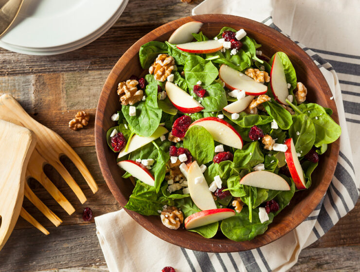 Fresh spinach salad with apple slices, walnuts, cranberries, and feta cheese in a wooden bowl on a rustic table.