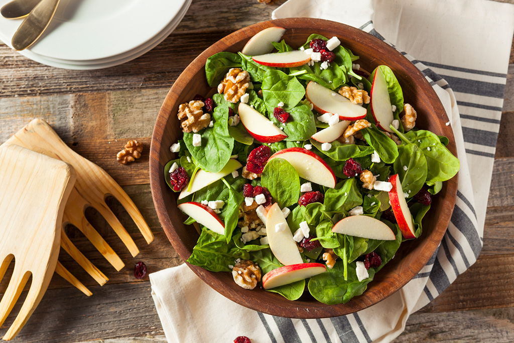 Fresh spinach salad with apple slices, walnuts, cranberries, and feta cheese in a wooden bowl on a rustic table.