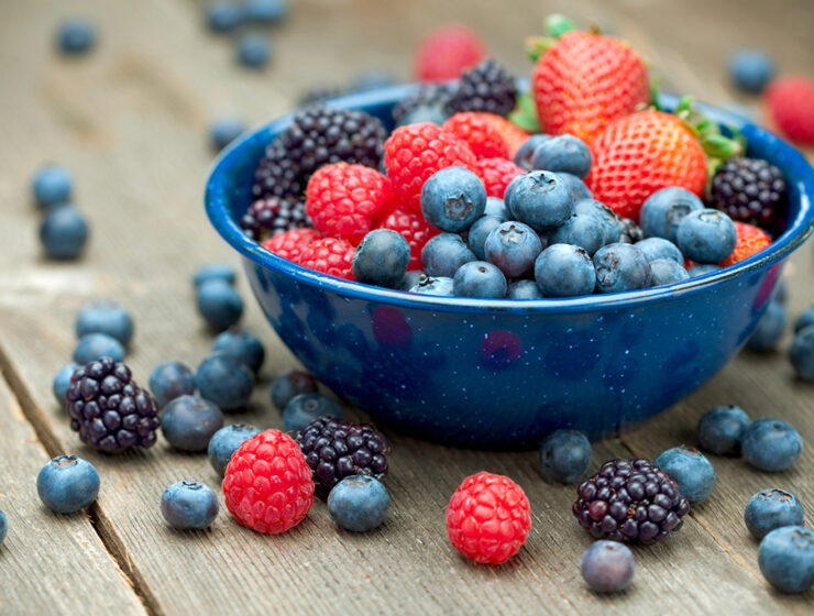 Assorted fresh berries in a blue bowl, including blueberries, raspberries, strawberries, and blackberries on a wooden surface for a healthy snack.