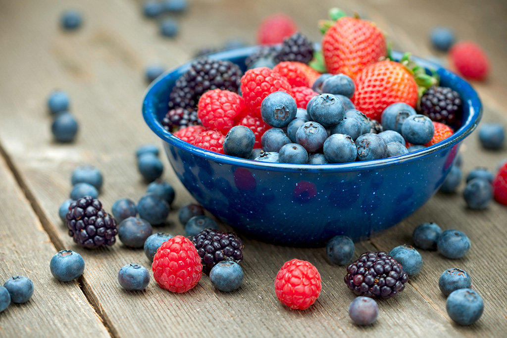 Assorted fresh berries in a blue bowl, including blueberries, raspberries, strawberries, and blackberries on a wooden surface for a healthy snack.