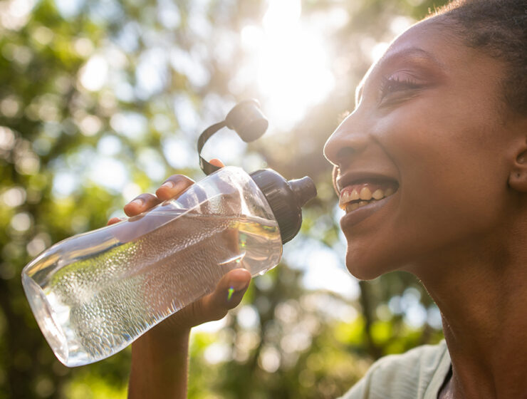 A person drinks from a clear water bottle, illuminated by sunlight, surrounded by lush greenery.