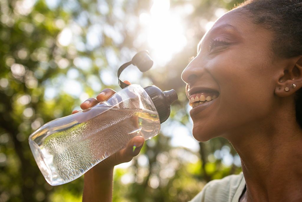 A person drinks from a clear water bottle, illuminated by sunlight, surrounded by lush greenery.