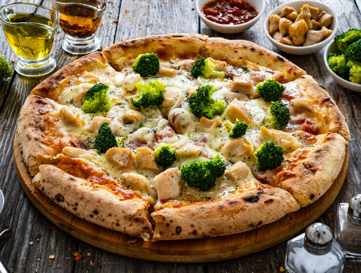Delicious pizza topped with broccoli and chicken on a wooden table, accompanied by olive oil, chili flakes, and side dishes.