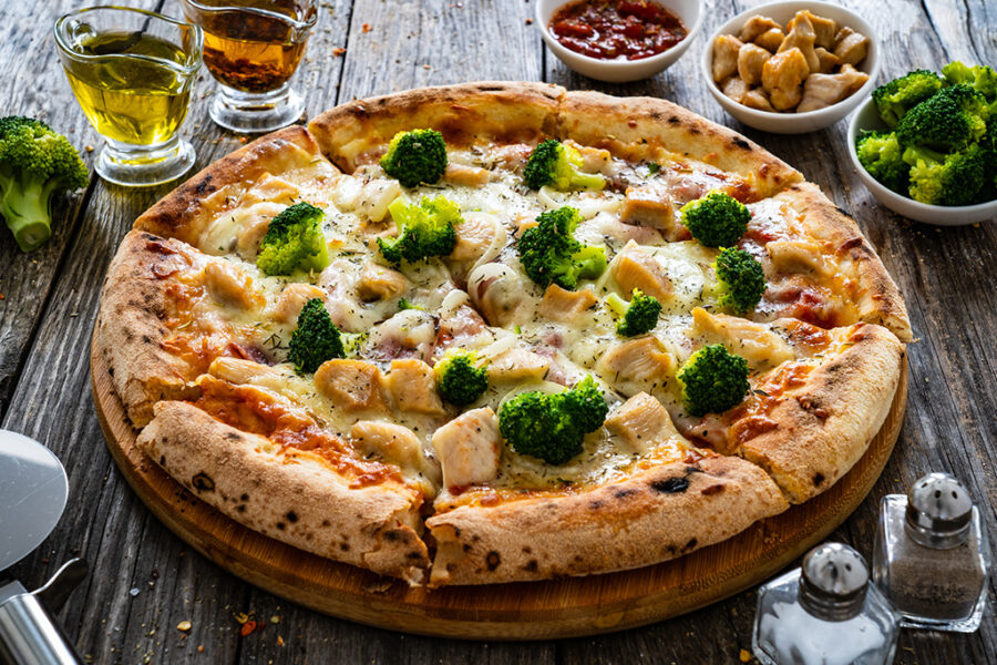 Delicious pizza topped with broccoli and chicken on a wooden table, accompanied by olive oil, chili flakes, and side dishes.
