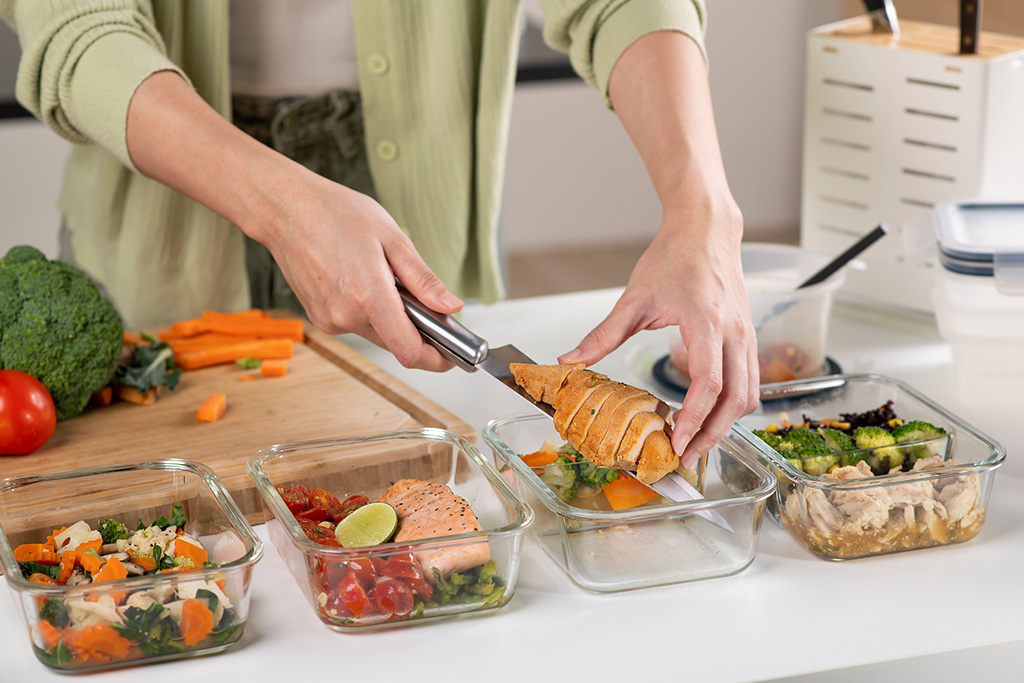 Person meal prepping healthy food with chicken, salmon, and vegetables in glass containers.